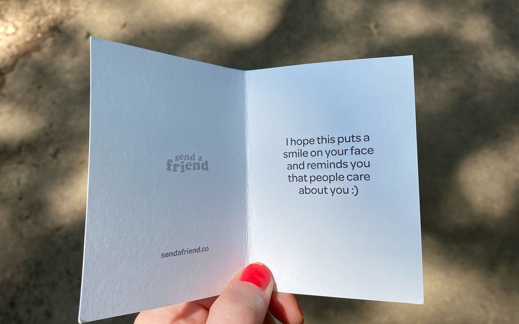 A picture of a hand holding an open SendAFriend note card that says "I hope this puts a smile on your face and reminds you that people care about you :)"