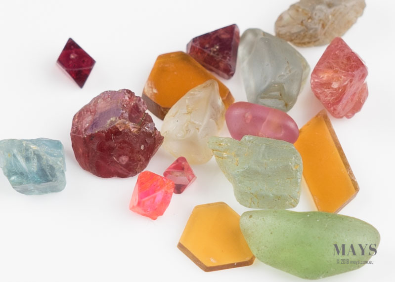 A mix of gem quality roughs with some in crystal form. In the mix are: Aquamarine, Ruby, Spinel crystals, Sapphire, Peridot, Topaz on a white background.