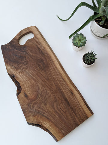 Walnut charcuterie board with grip handle with white background.  Succulents in white pots in the background. 