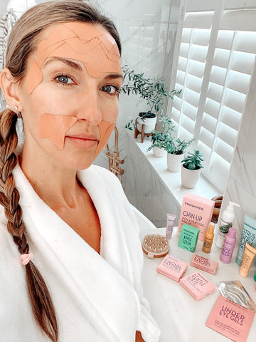 woman with braid wearing Frownies Facial Patches on forehead, corners of the eyes, and corners of the mouth with Frownies skincare products on the counter behind her