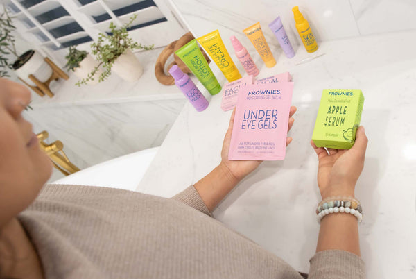 over the shoulder of woman holding pink package Frownies Under Eye Gels and green box of Frownies Apple Serum, with other Frownies skincare products on bathroom counter