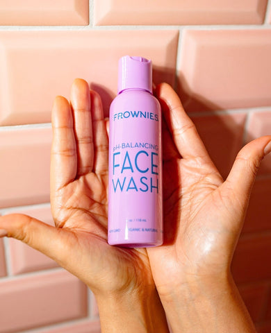 hands holding Frownies pH Balancing Face Wash purple bottle against pink tile background