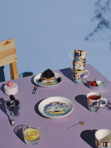 Der Mumin Mittsommer-Becher 2024 Berry Season auf der gedeckten Tafel: Mit dem passenden Teller und Gläsern, Törtchen und Himbeeren. Und das ganze farblich passend auf einer flieder-farbenen Tischdecke.