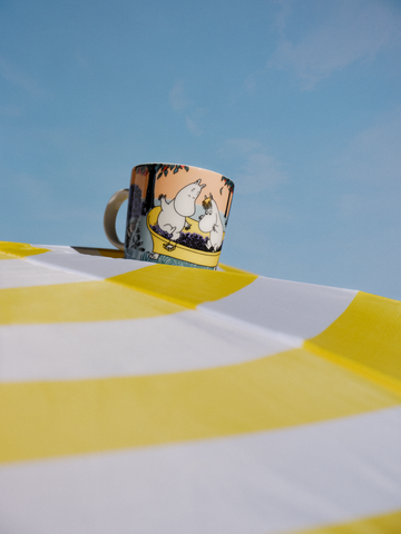 Die Mumin-Mittsommer-Tasse 2024 Berry Season/ Marjassa bei HARTOG: Moomin und Snorkfräulein stampfen Beeren in der Badewanne. Die Tasse steht auf einem gelb-weiß gestreiften Badetuch vor blauem Himmel.