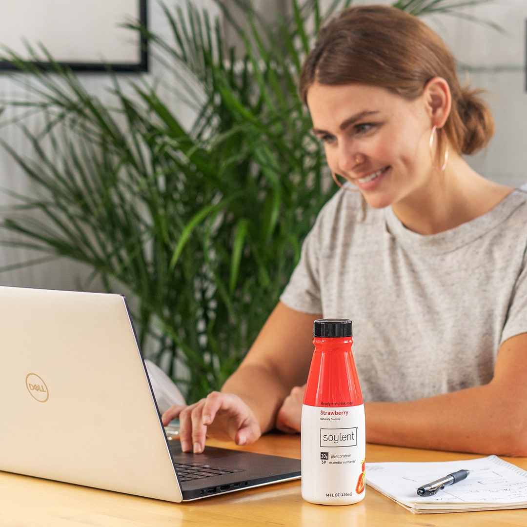Image of woman working at computer with a bottle of soylent next to her