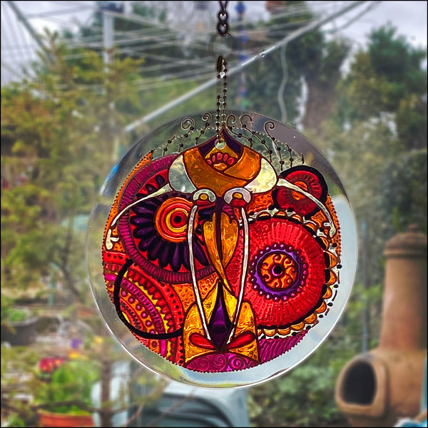 Stained glass suncatcher hand painted with an intricate red pink mandala design, hanging in a window looking out on a garden