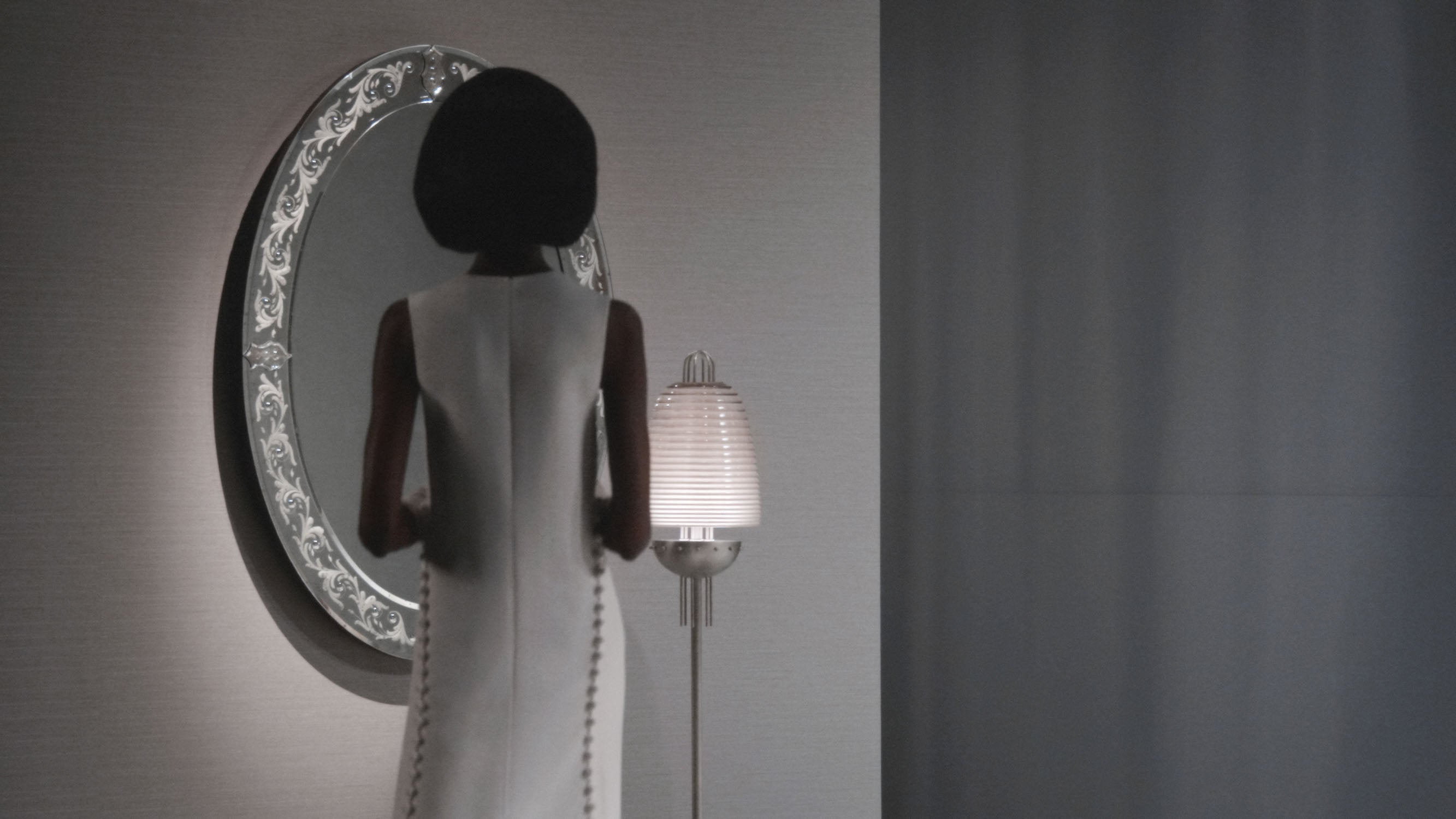 A woman stands in front of an oval mirror, next to a SIGNAL : X floor lamp. 