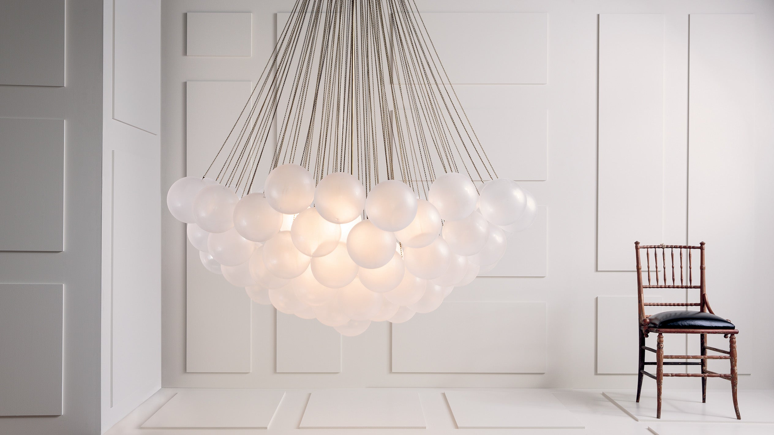 An illuminated CLOUD : XL 73 chandelier in Aged Brass finish, hanging next to a chair in a white paneled room.