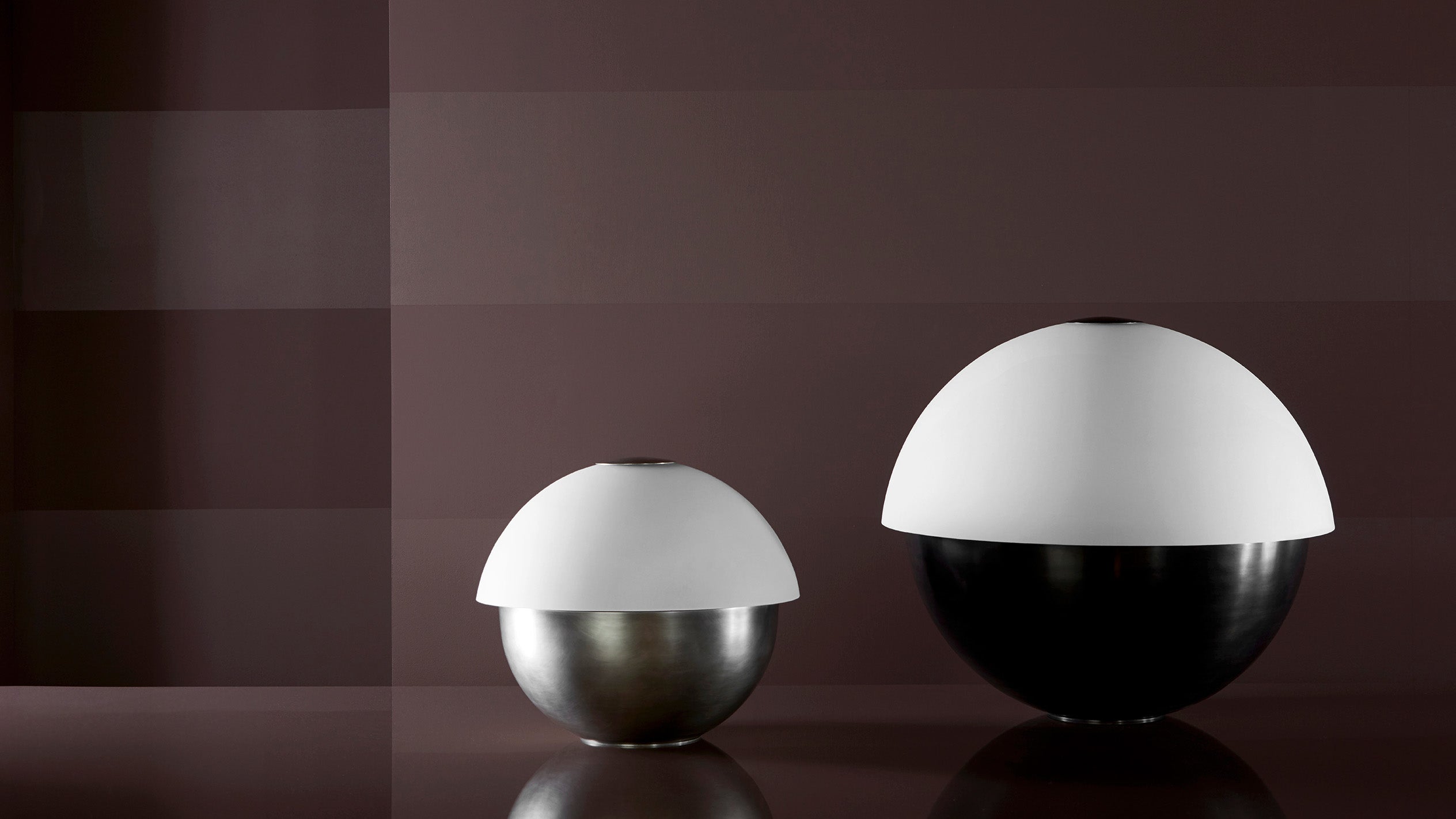 AXON : SMALL table lamp in Tarnished Silver and AXON : LARGE table lamp in Blackened Brass on a surface.