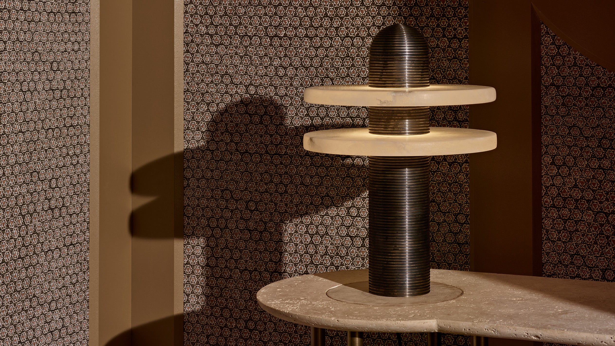 An illuminated MEDIAN table lamp is sitting on a console table in front of decorative wallpaper. 