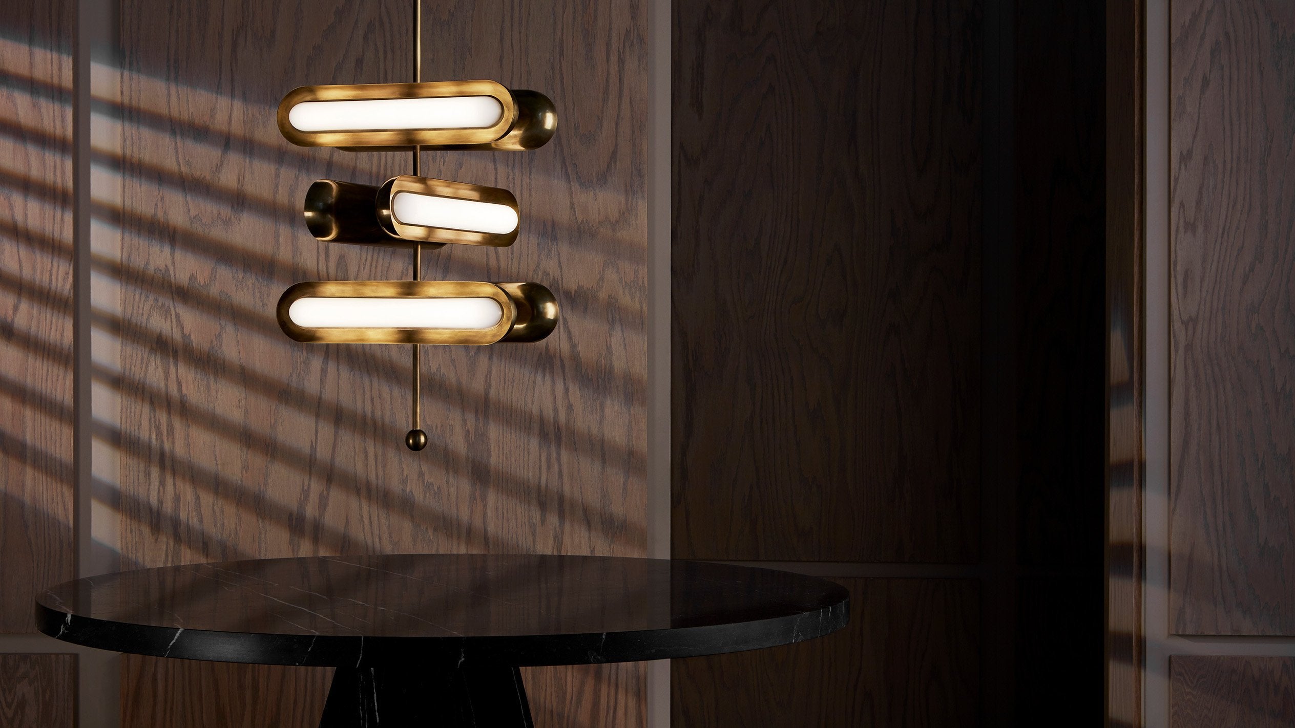 CIRCUIT : 6 ceiling pendant in Aged Brass finish, hanging above a table with natural light cast on the wall behind it. 