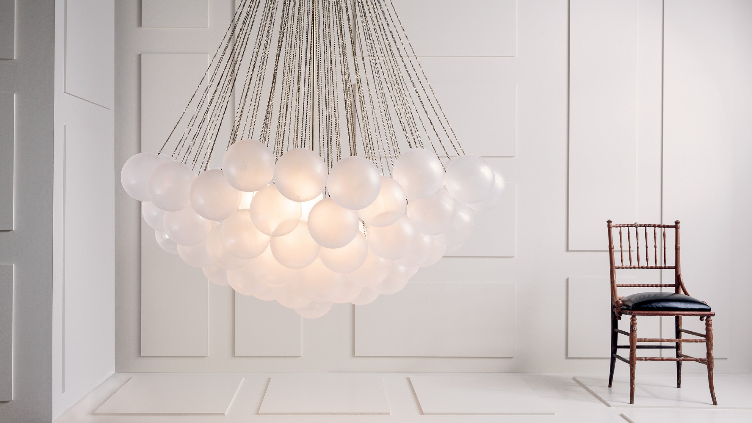 An illuminated CLOUD : XL 73 chandelier in Aged Brass finish, hanging next to a chair in a white paneled room.