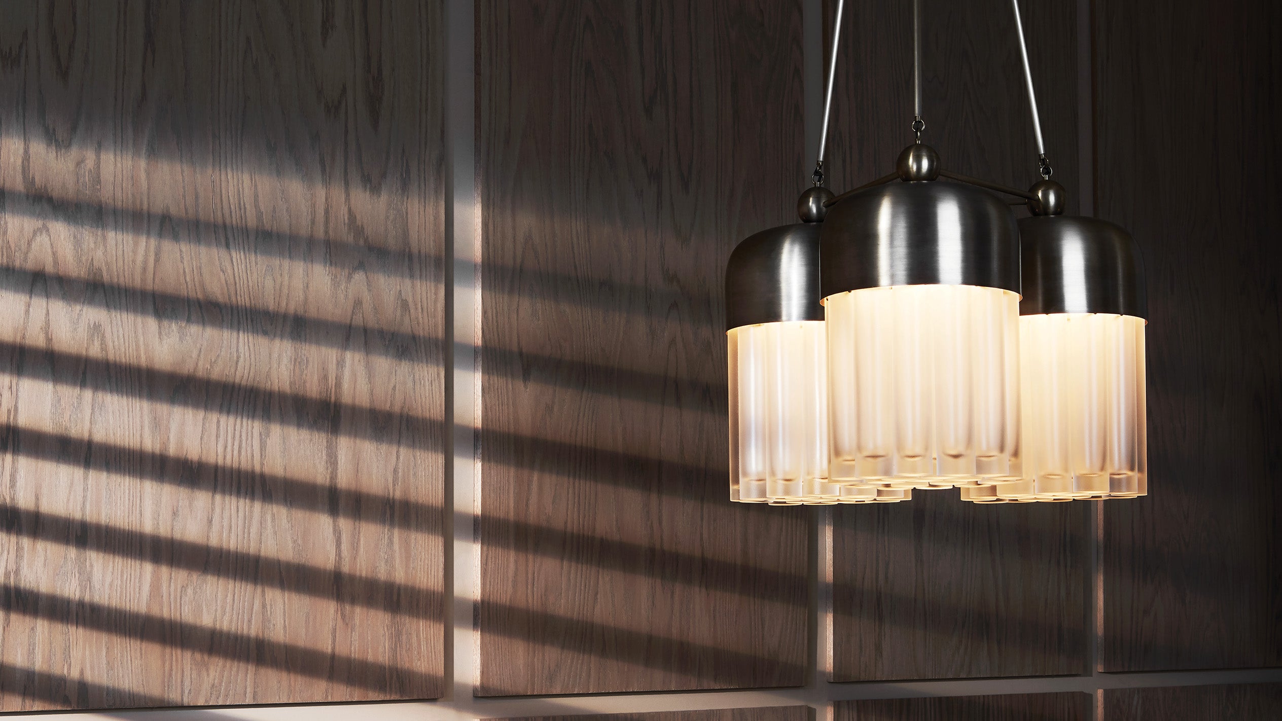 An illuminated TASSEL : 57 chandelier hanging against a wood textured wall. 