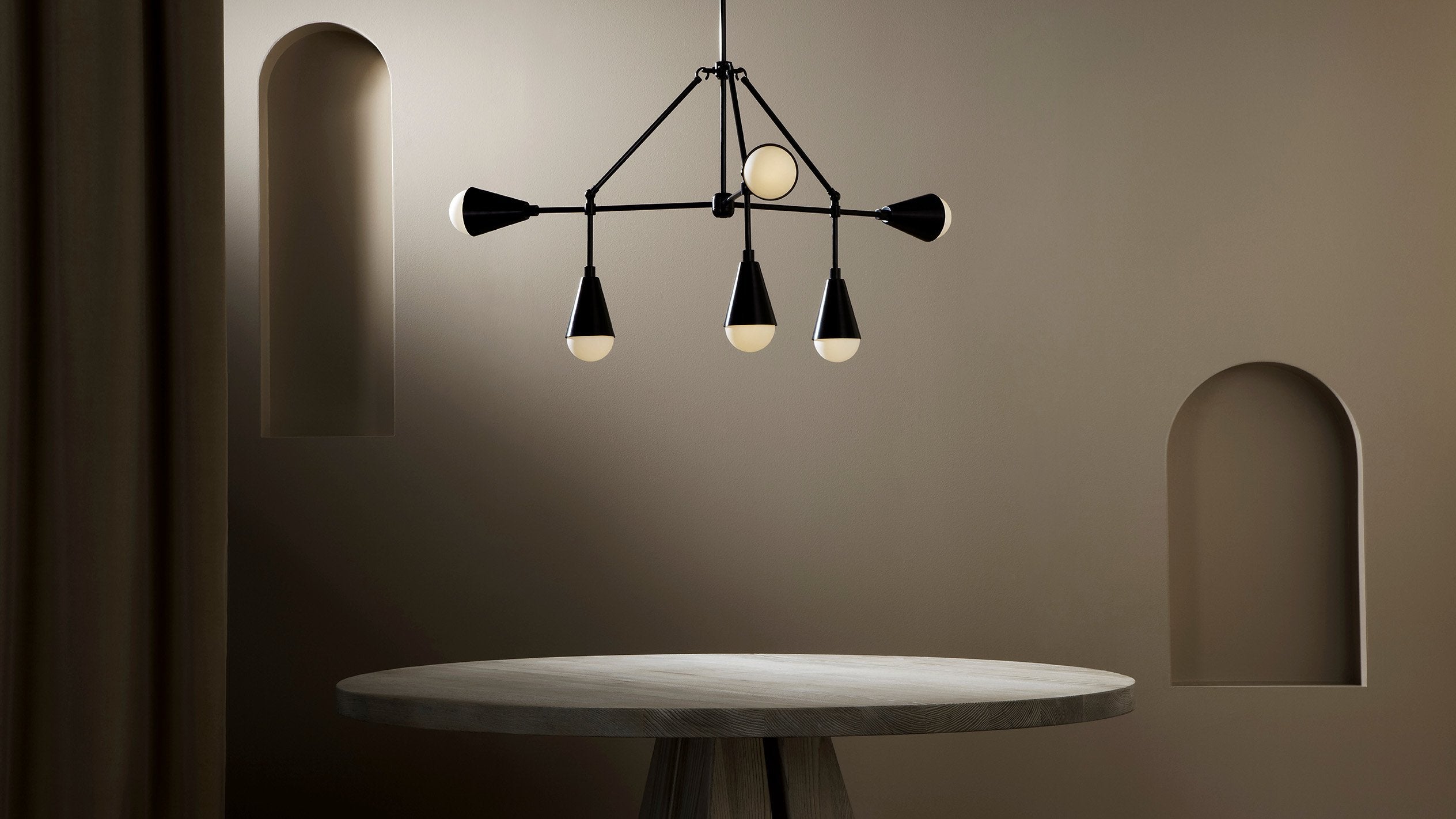 An illuminated TRIAD : 6 ceiling pendant in Blackened Brass finish hanging over a circular dining table. 