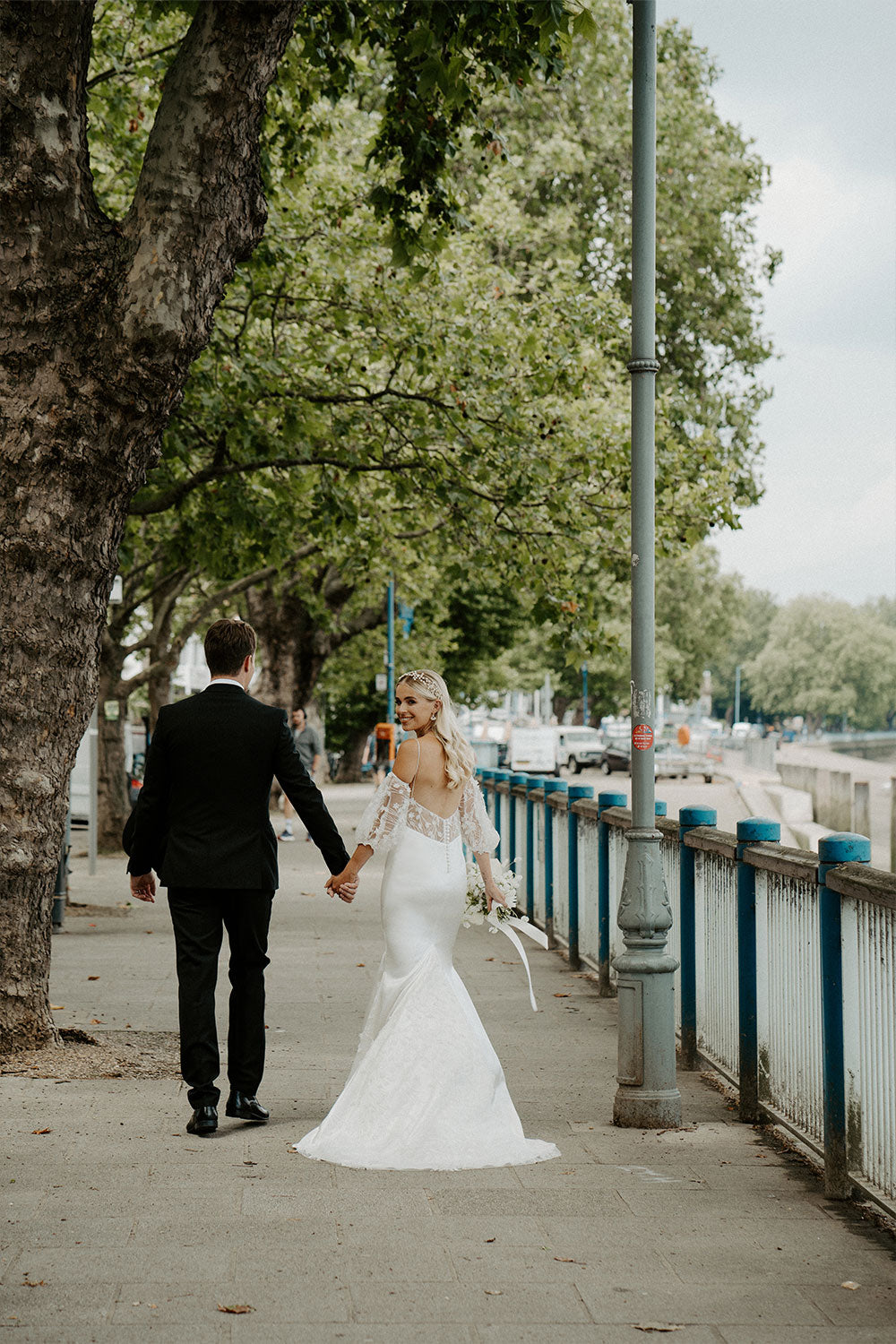 Bride and groom walking along Putney Embankment. The bride is wearing teh Florence Wedding Dress designed and made by Constellation Ame in London.