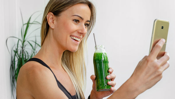 woman taking a selfie of her holding a green drink in a glass bottle