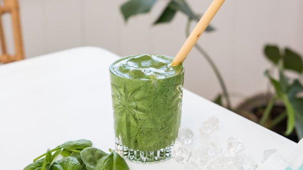 green smoothie in a glass with a bamboo straw sitting on a white table with loose spinach in front