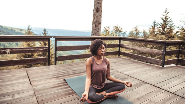 A woman sitting in a cross-legged meditation position on a yoga mat. She is outdoors on a timber balcony and there is a background of hills and nature. She is wearing a brown tank top and black yoga pants. She has chin-length dark brown curly hair.