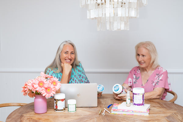 Amanda and Robyn are sitting at a timber dining table in front of a macbook laptop and four of their products. They are in floral dresses and smiling