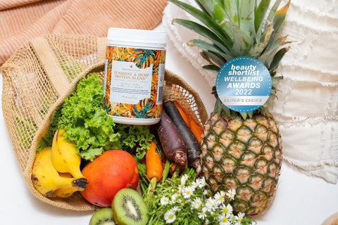 Myella Sunshine & Hemp Protein blend in a basket with assorted fruits including pineapple, black and orange carrots, bananas, kiwifruit, kale and mango - all found in this nutritional blend