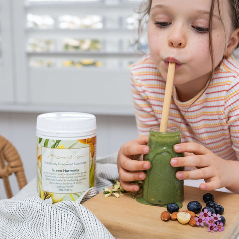 Young girl leaning on the kitchen bench sipping on a green smoothie in a glass jar through a bamboo straw. A container of Mayella Green Harmony nutritional blend used to make the smoothie is on the bench next to her.