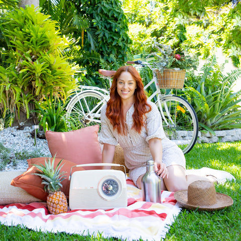 Garden setting with a woman sitting on picnic blanket with pineapple, Mayella stainless steel drink flask, vintage radio and bicycle in background.