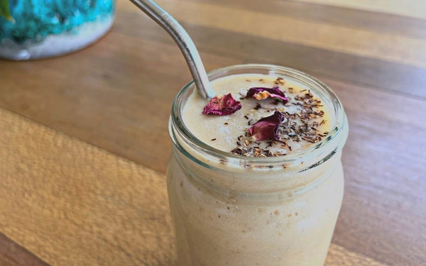 A glass jar filled with a beige-coloured smoothie, topped with a sprinkle of loose rooibos tea and dried, red rose petals. It's on a timber table with a bluey-turquoise pot slightly showing in the background.
