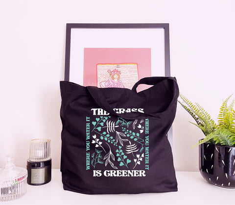 A black tote bag with a floral design that reads The Grass Is Always Greener Where You Water It