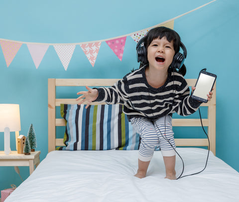 Child listening to music via a phone and headphones on a bed.