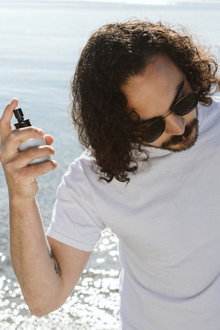 man spraying hair at the beach with sea salt spray in a glass bottle