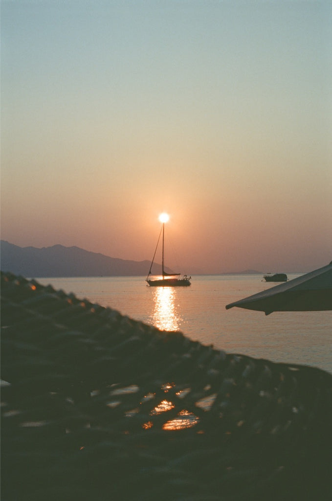 Sunset over calm waters, a sailboat silhouetted with its mast aligned to the rising sun, reflecting on the sea. Captured on Kodak Ektachrome 100 35mm film.