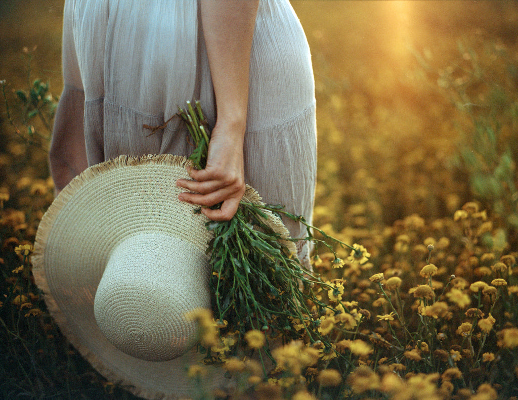 a women standing in a field of yellow flowers holding a hat at her waist.