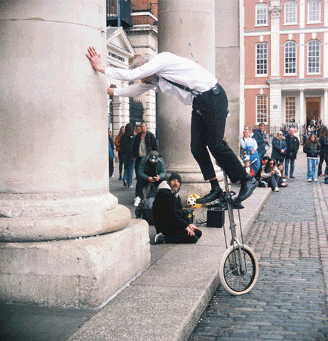 A stereoscopic GIF of a street performer balancing on a unicycle, leaning against a large column, with an audience watching in a plaza.