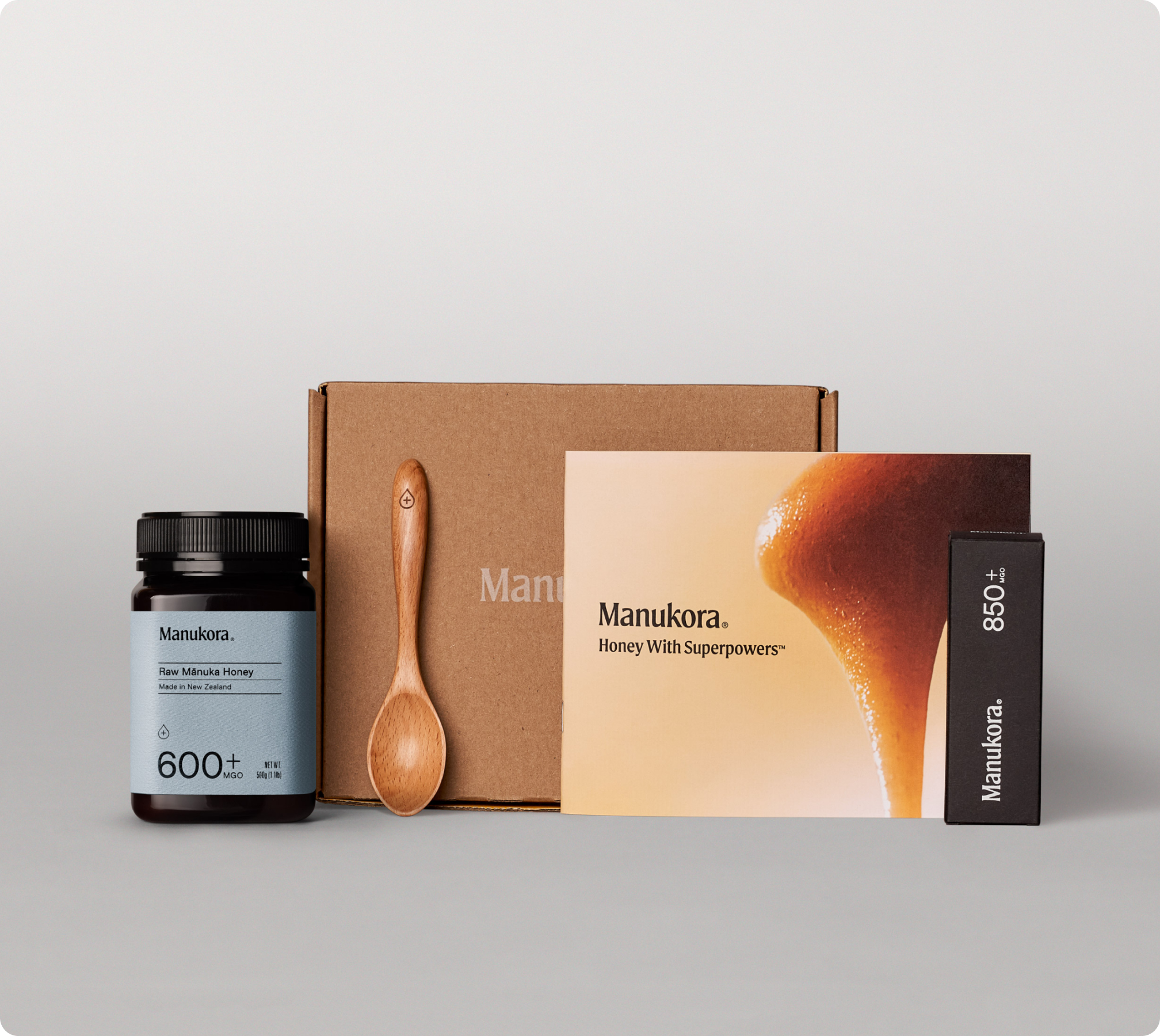 Manukora honey products, wooden spoon, and packaging box displayed on a plain background.