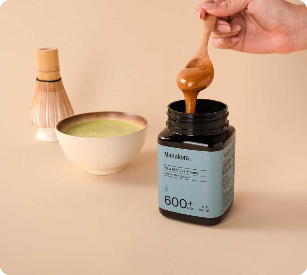 A hand dipping a spoon into a jar of Manuka honey next to a bowl of green matcha tea.