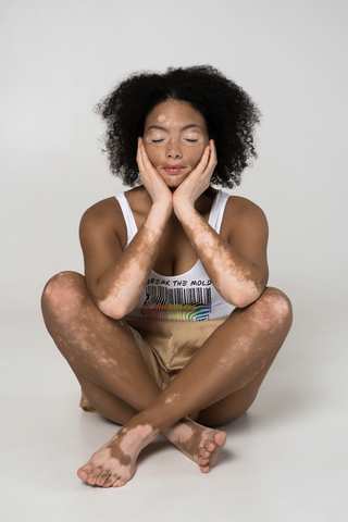 A Black model with vitiligo is sitting cross-legged on the floor wearing shorts and a white short-sleeve Break the Mold shirt. In the middle of the tee is a graphic of zebra in the silhouette of a barcode. The top half of the zebra are vertical black stripes, which then change just below the back into the colors of the rainbow. The bottom of each stripe has a paint drip mark. Just above the zebra's back is the phrase, in upper case, 'break the mold.'