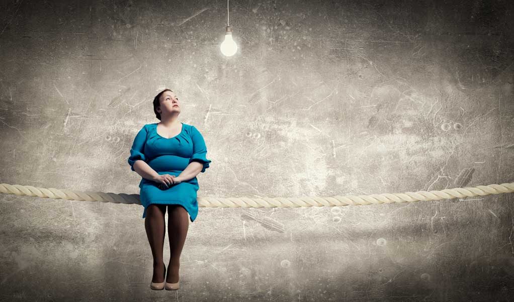 A person living with scoliosis in a blue dress is sitting on a rope looking intently at a light bulb.