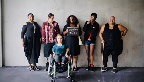 A diverse group of disabled people standing together, smiling confidently in front of a concrete wall. Some use mobility aids, and one holds a sign reading “disabled and here.”