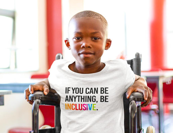 This is a color photo on the classic Be Inclusive kids t-shirt by URevolution. A child is sitting in a wheelchair wearing a white t-shirt with the phrase, 'If you can be anything, Be Inclusive,' printed on it in upper case black letters. The word inclusive is in the colors of the rainbow.