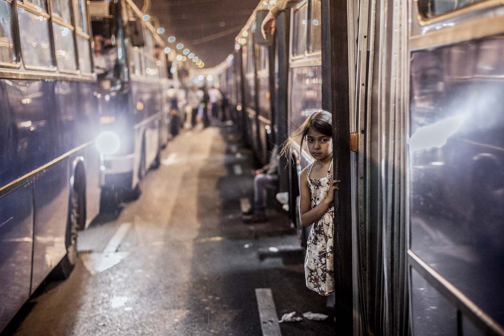 A photo of a migrant girl standing in the entrance of a bus at night. The bus is in a row of buses, lined up next another row of buses.