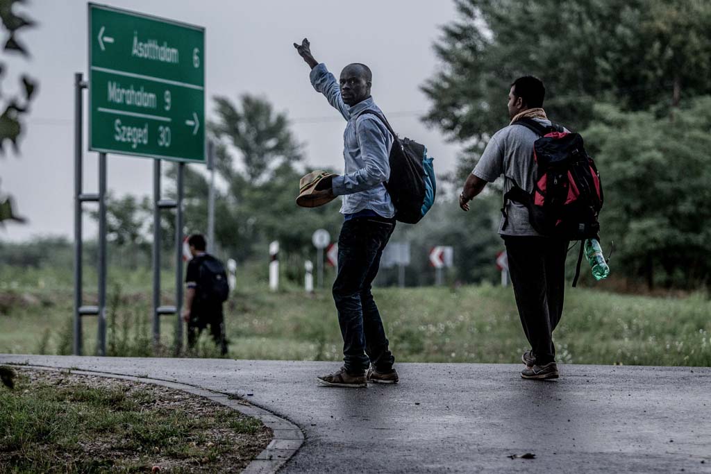 Two migrants are walking on a road. Behind them is a sign giving directions to a number of cities. Being on the move, in the countryside, gives rise to many migrant health issues.