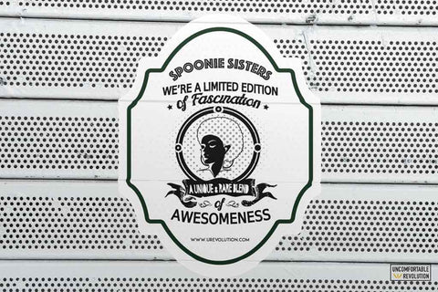 A photo of kiss cut spoonie sister sticker on a outside metal wall. The sticker has black writing on a white background. 1st line: "SPOONIE SISTERS." 2nd line "WE'RE A LIMITED EDITION." 3rd line "of Fascination." In the middle of the sticker is a roundel with a spoon sister with an afro. On the 5th line, on a banner under the roundel, "A UNIQUE & RARE BLEND." 6th line "of." 7th line "AWESOMENESS." "www.urevolution.com" is printed in small font at the bottom of the sticker.