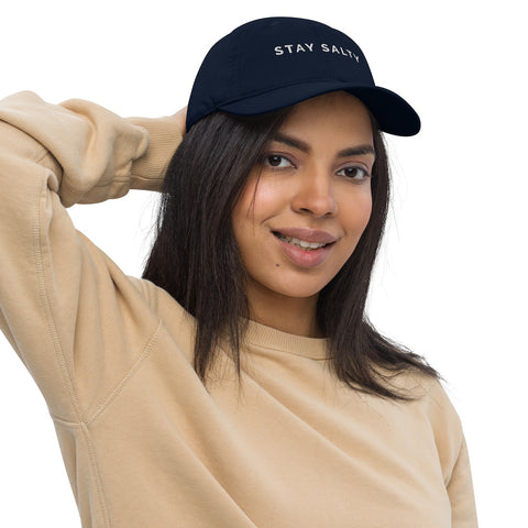 A photo of a navy Stay Salty cap worn by a model with long black hair and a brown sweatshirt. On the front panel of the cap, the phrase, 'Stay Salty,' is embroidered in white upper case letters.