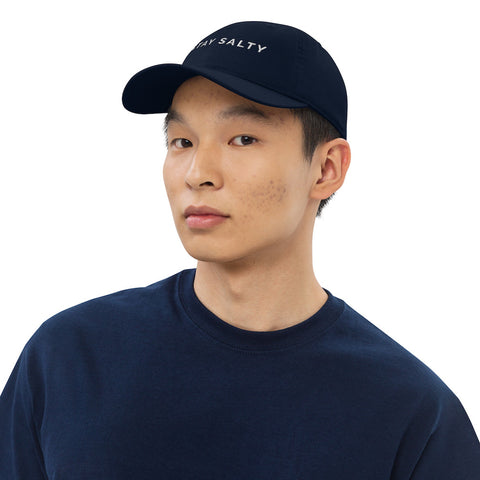 A photo of a navy Stay Salty cap worn by a model with short black hair and a navy sweatshirt. On the front panel of the cap, the phrase, 'Stay Salty,' is embroidered in white upper case letters.