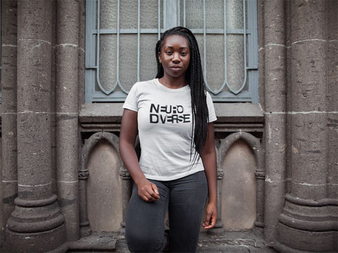 A woman is standing against a stone building wall wearing a white neurodiverse shirt. The neurodiverse shirt she is wearing has the text NEURODIVERSE printed in large alternating black and white letters, taking up about one-third of the t-shirt surface.