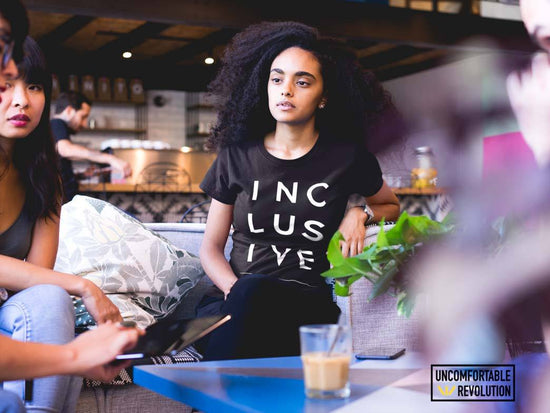 Beneficios de la inteligencia emocional en el ámbito laboral en una cafetería.  Los compañeros de trabajo están hablando entre sí.  La persona en el centro tiene el pelo largo y ondulado y lleva una camiseta negra con la palabra inclusiva impresa.