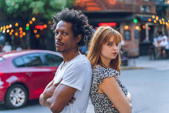 An annoyed mixed-race couple is standing back to back on the street with crossed arms trying to develop connections despite being in great physical pain.
