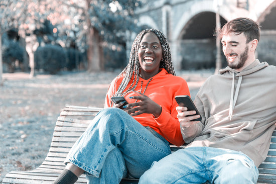 Feliz pareja joven multiétnica mirando un teléfono móvil en el parque sentado en un banco riéndose de respuestas divertidas a ¿Cómo estás?