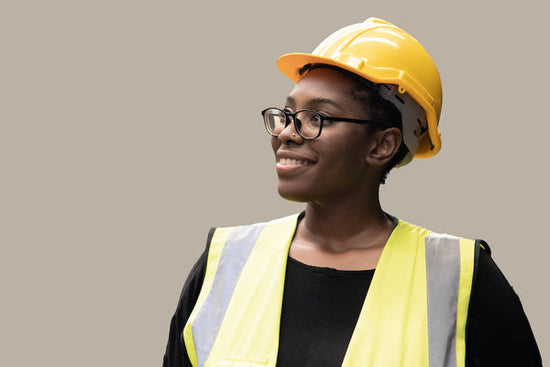 Mental well-being as a manager is essential for productivity: a Black woman is standing against a wall wearing a yellow safety vest and safety helmet.