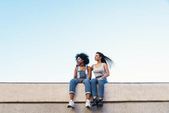 Two women sit on what looks like the top of a concrete road bridge. The both look off defiantly to the left The woman to the right has her arm around the other. They are both silent, and not talking. They are casually dressed in jeans and tank tops.